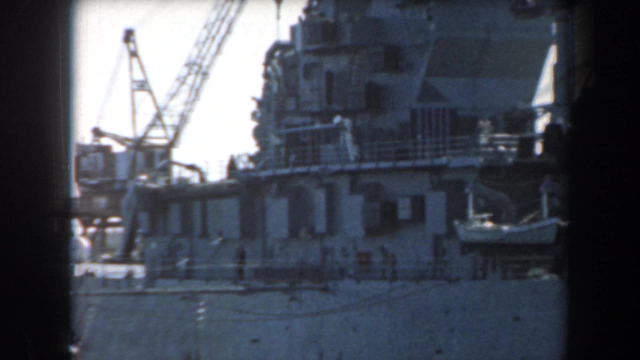 Large warship beside a dock with cranes rising behind it.