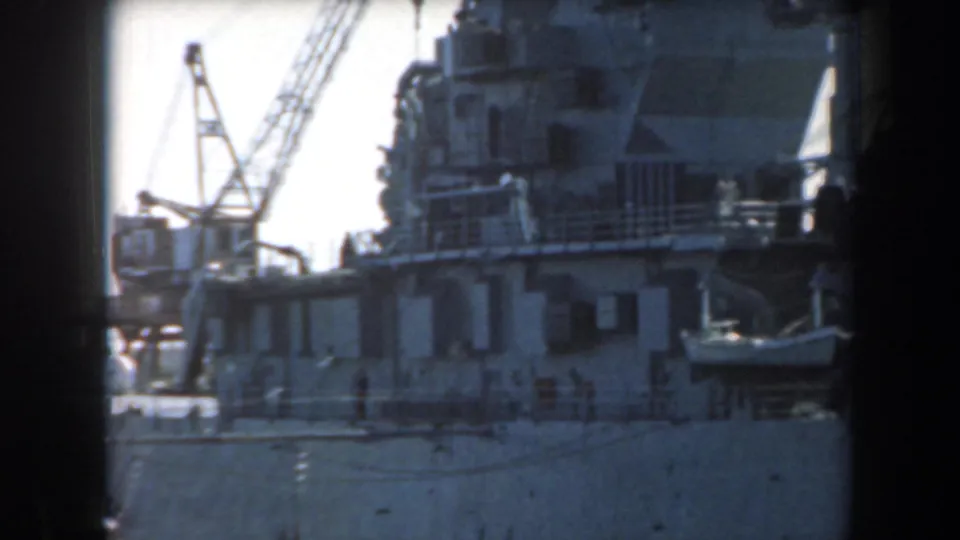 1961 California home-movie frame showing a large naval vessel near dockside cranes in a harbor setting.