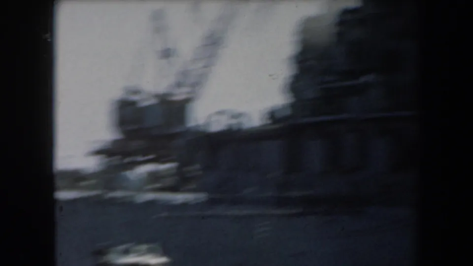 Motion-blurred 1961 home-movie frame panning past a naval ship and port crane at a California harbor.
