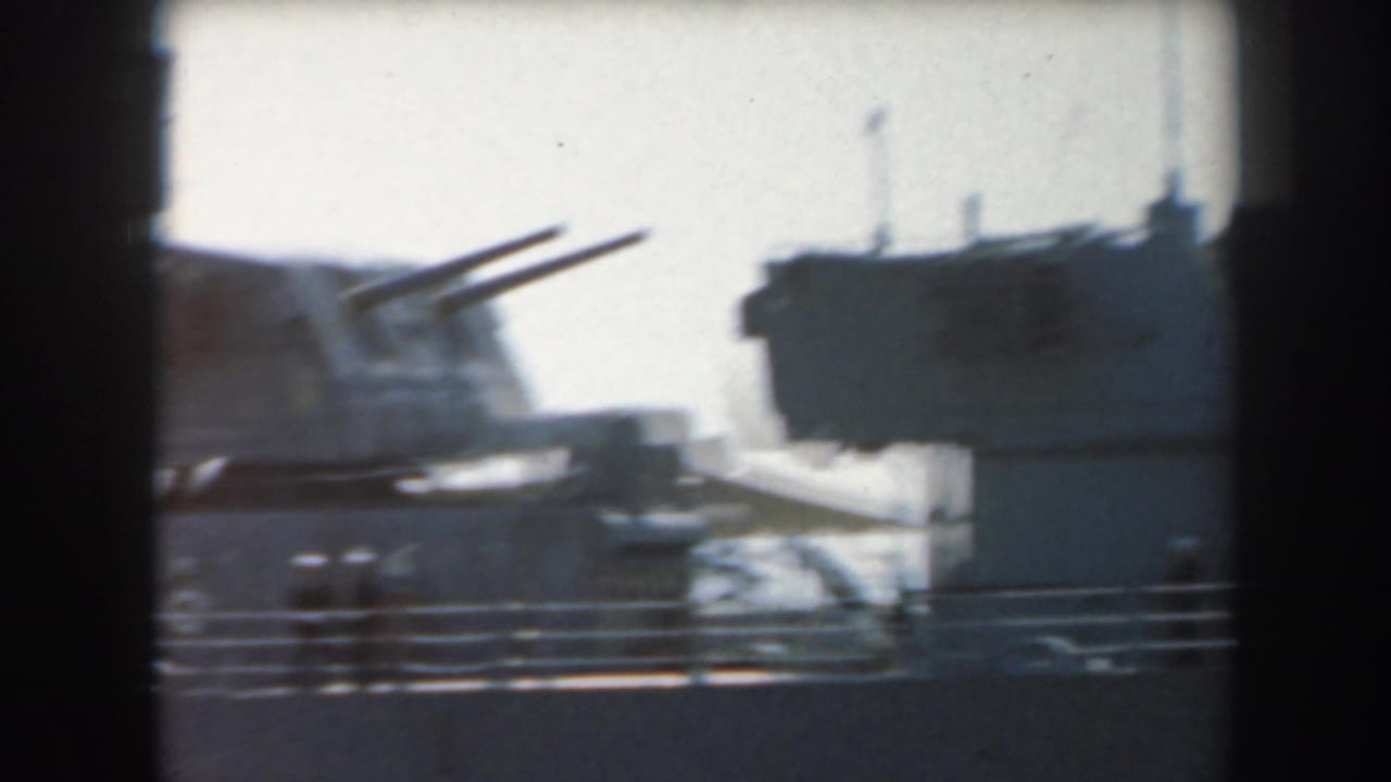 Warship deck with twin naval guns and a boxy superstructure seen through dark film borders.