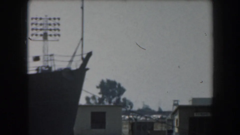 The ship's bow and rigging appear in silhouette beside harbor buildings and a tall light tower in the 1961 California home-movie footage.