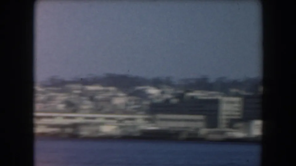 A distant California shoreline with waterfront buildings, blue water, and low hills closes the brief 1961 harbor view.