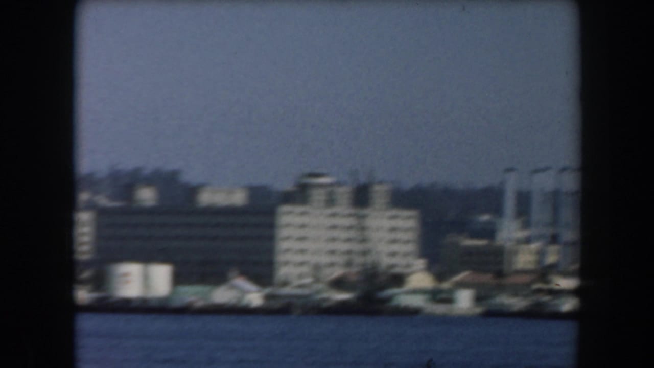 1961 California home-movie frame showing white and grey waterfront buildings across blue harbor water.