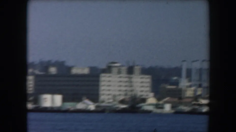 1961 California home-movie frame showing white and grey waterfront buildings across blue harbor water.