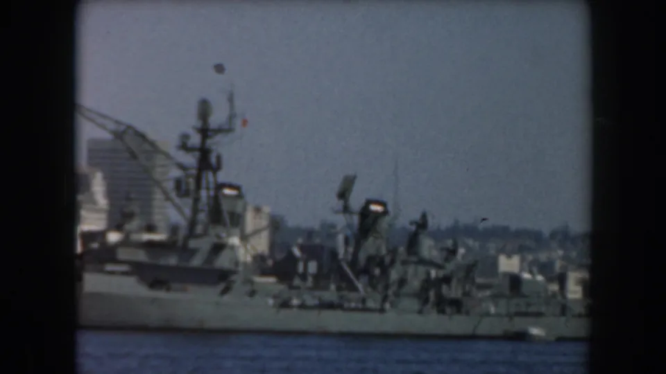 Grey naval-looking ship on blue harbor water with masts, equipment, and a hazy California skyline behind it.