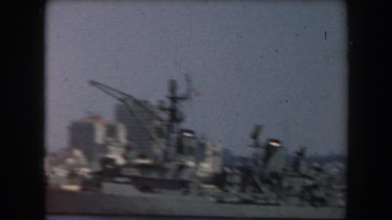 1961 California home-movie frame showing a grey ship near a hazy waterfront skyline from a moving vessel.