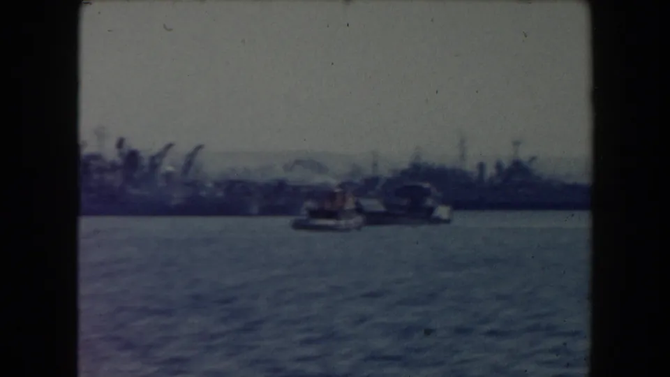 1961 California 8mm view across water toward a distant harbor with boats, cranes, and low shoreline structures.