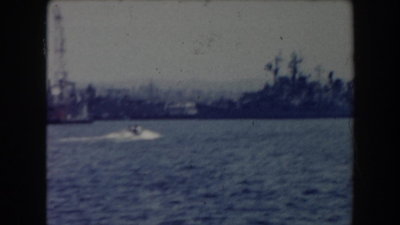 Distant docked ships across blue-grey harbor water with a small motorboat wake in front.
