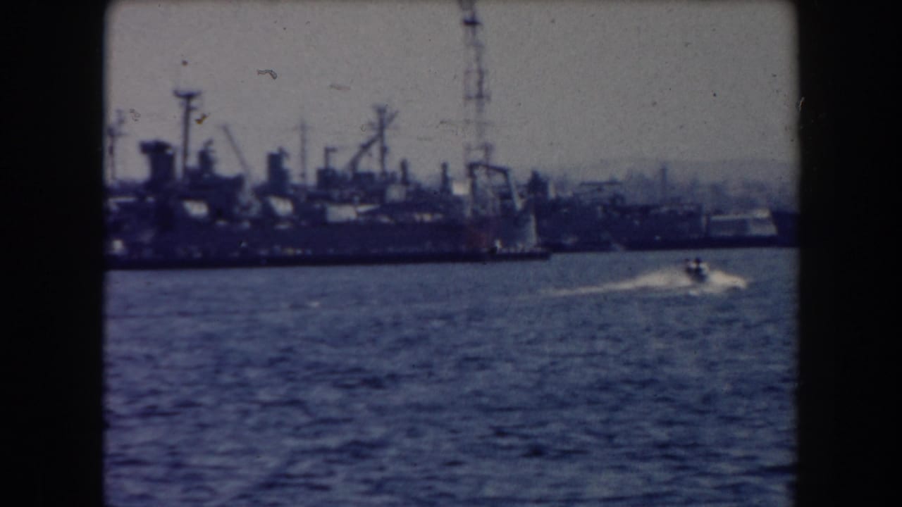 Large ships and cranes line the harbor as a small boat speeds across the water.