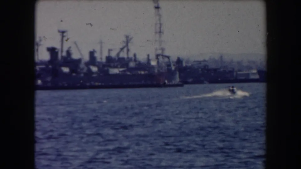 California port scene from 1961 with docked industrial vessels, cranes, and a small motorboat leaving a wake.