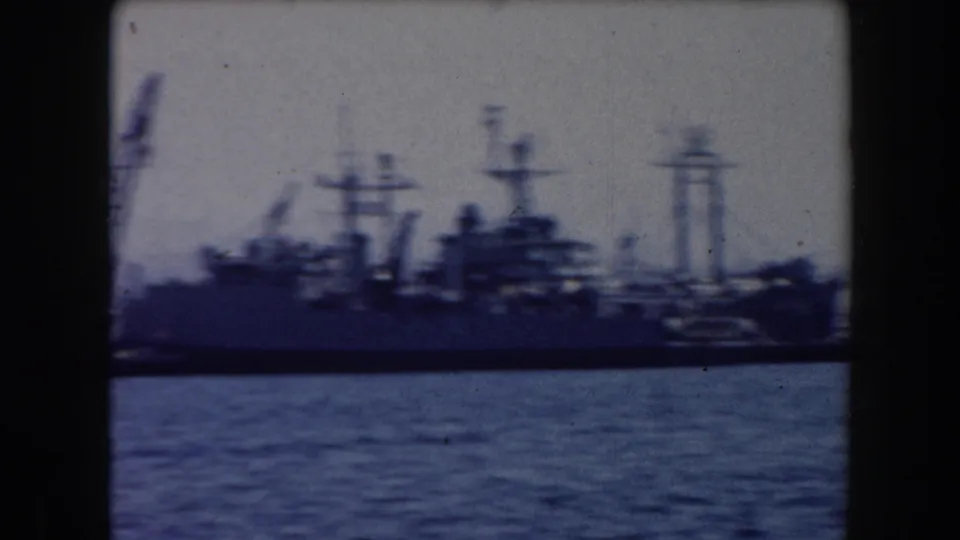 1961 California harbor footage showing a large grey docked ship with masts and waterfront structures beyond blue-grey water.
