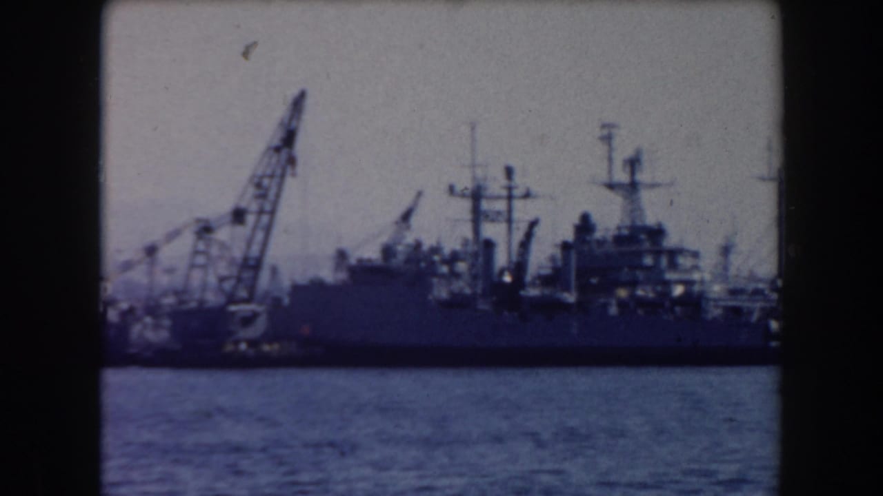 1961 California home-movie frame showing naval ships moored near industrial cranes across blue harbor water.