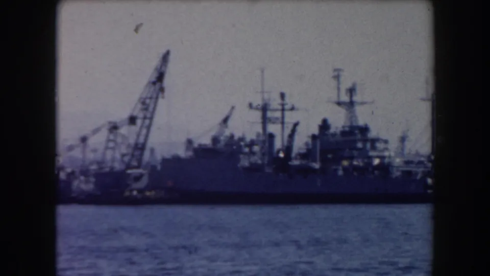 1961 California home-movie frame showing naval ships moored near industrial cranes across blue harbor water.