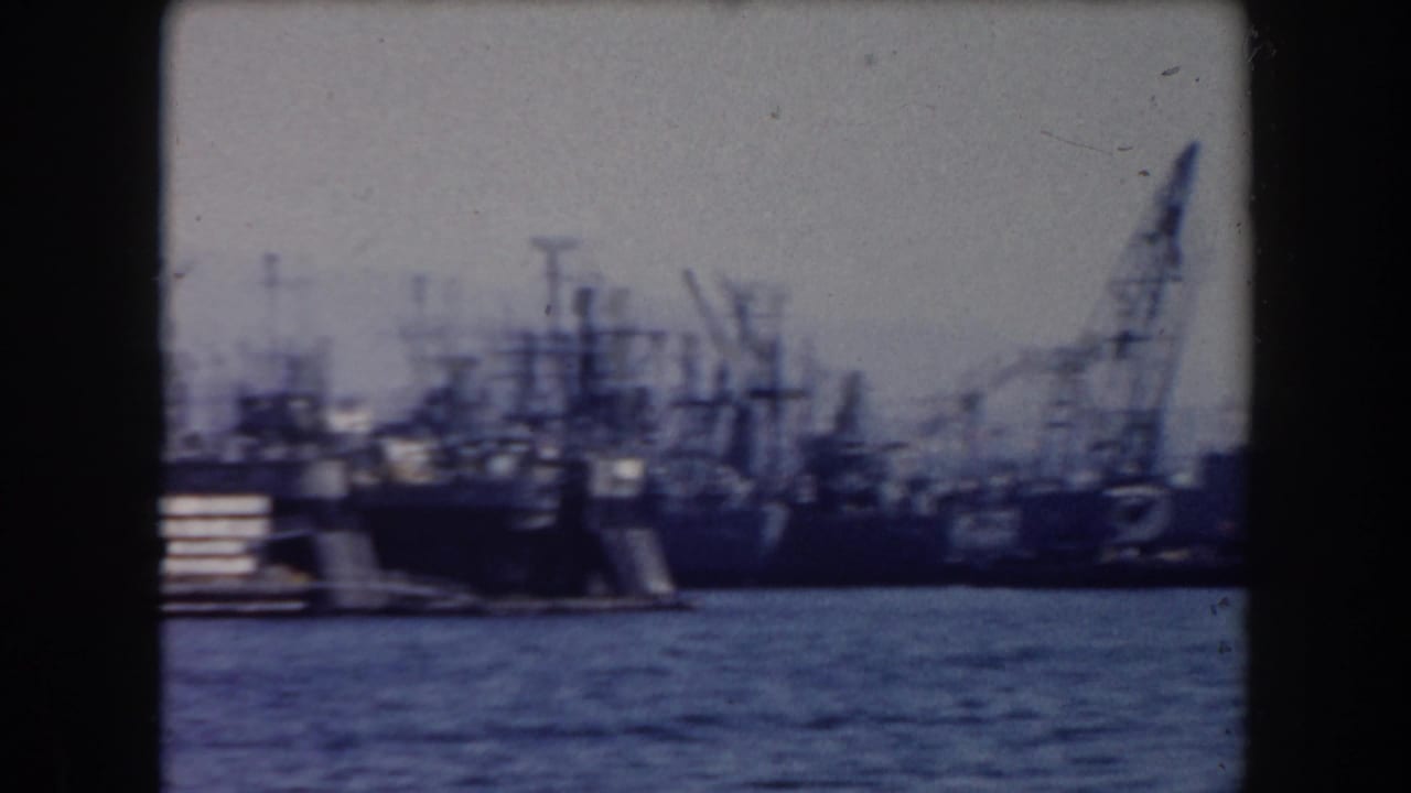 Warships moored near cranes with water in foreground.