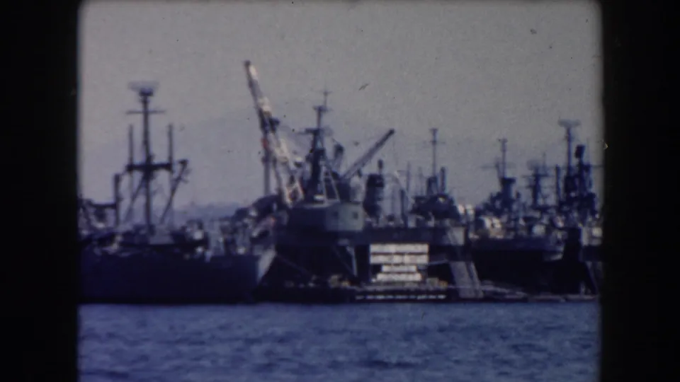 A harbor crane and several moored naval ships appear in soft, grainy 8mm footage from the 1961 California clip.