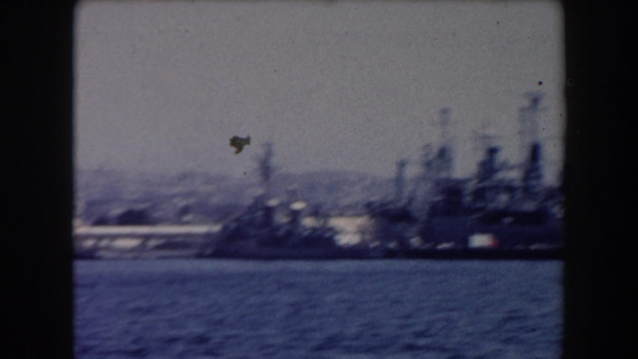 Distant gray warships anchored across calm harbor water with hills behind them.