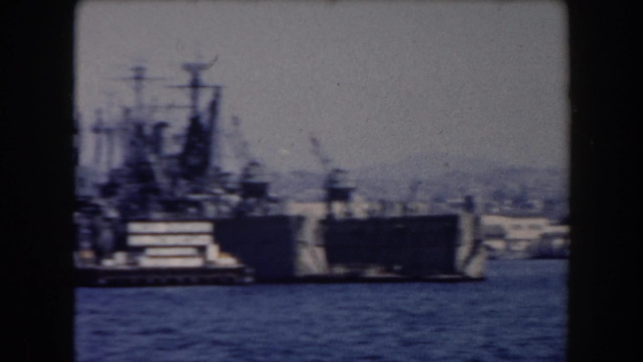 Large naval vessel near a pier with another ship and harbor shoreline in the background.