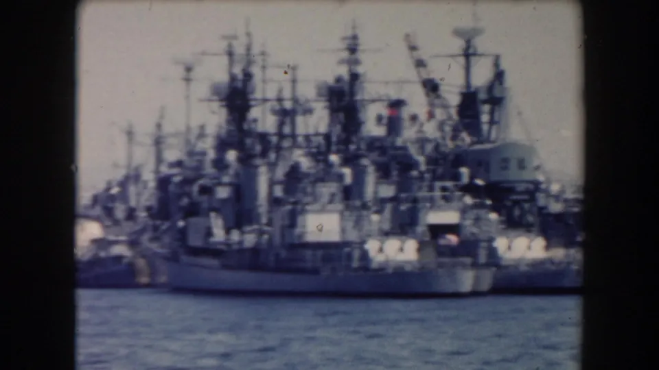 The pan settles on docked gray warships with tall masts and layered superstructures in a 1961 California harbor setting.