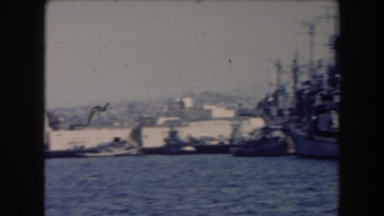 1961 California harbor view with docked naval vessels, blue water, low buildings, and dark 8mm frame borders.