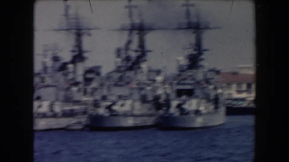 1961 California harbor frame showing several docked naval ships with tall masts and superstructures near waterfront buildings.