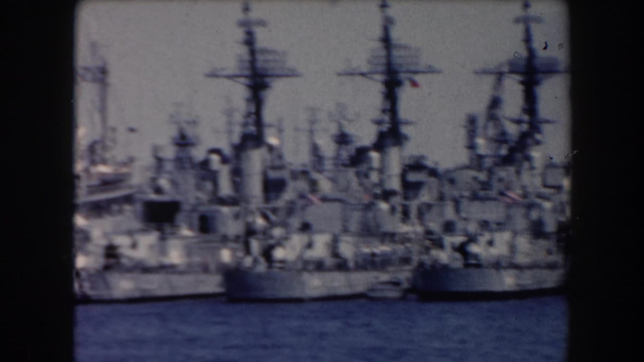 Several docked naval vessels with tall masts seen across harbor water.