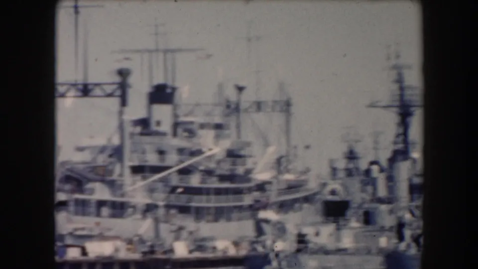 California port scene with naval-looking vessels, dockside structures, cranes, and industrial harbor equipment visible in soft 8mm focus.