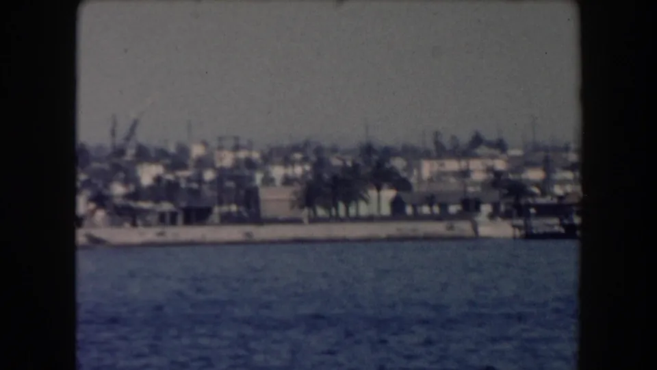 Wide 1961 California harbor view over water toward a low shoreline with buildings, trees, and distant port activity.