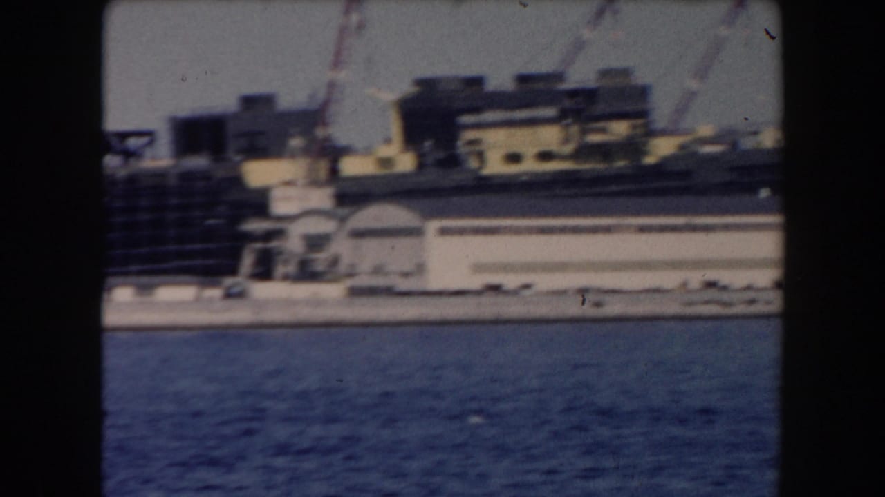 1961 California harbor view from water with docked vessels, low waterfront buildings, and cranes in soft 8mm focus.