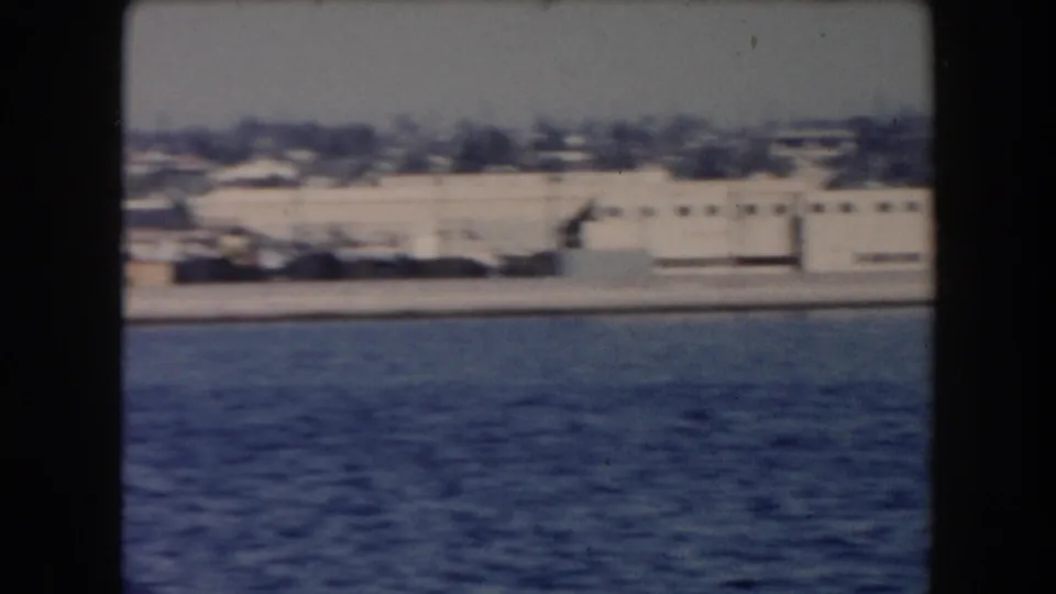 California harbor shoreline in 1961, seen across blue water with low industrial buildings and a hazy background.