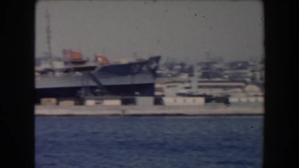 A large gray naval-looking ship appears docked along a California waterfront, with cranes and harbor buildings behind it.