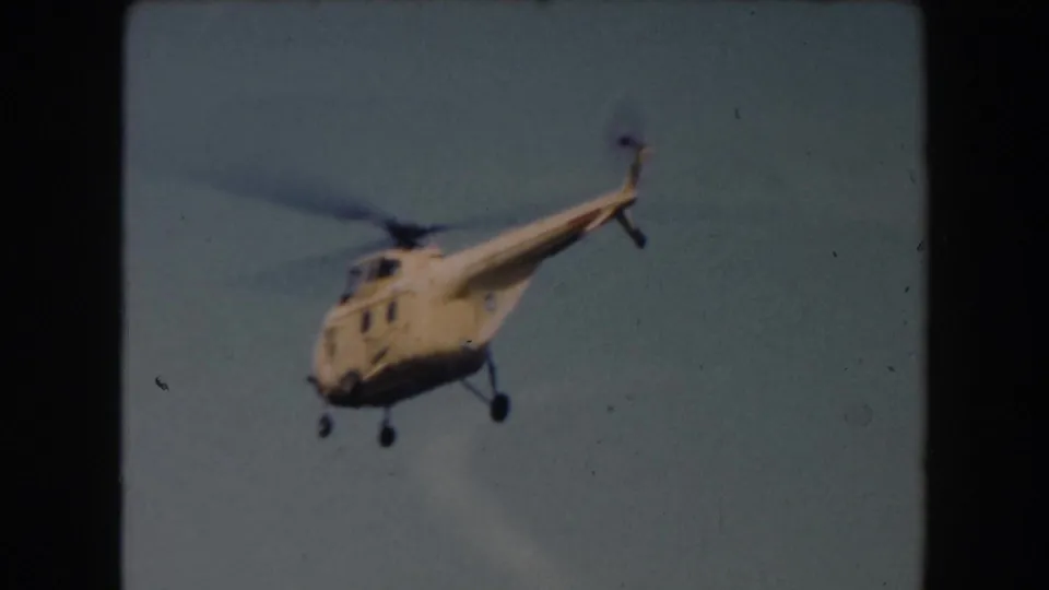 1961 California home-movie frame showing a yellow helicopter flying across a clear, pale sky.