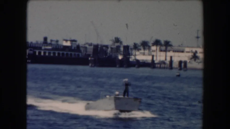 1961 California waterfront frame showing a small boat with a standing person, open water, a larger ship, harbor structures, and palm trees.