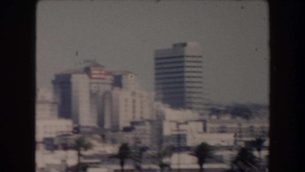 Soft city skyline view with a pale building, taller tower, palms, and shoreline structures.