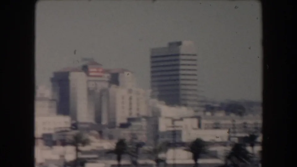 1961 California waterfront skyline seen through soft 8mm focus, with commercial buildings, palm trees, and a tall striped tower.