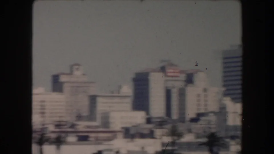 1961 California city pan showing blurred waterfront blocks, roadside cars, palms, and taller buildings under haze.