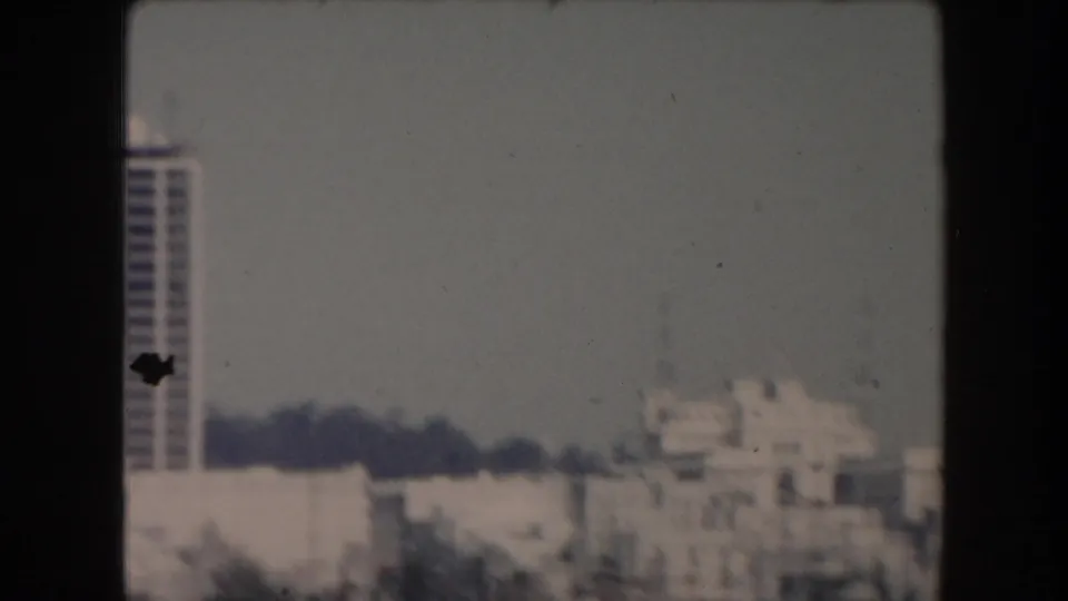 1961 California home-movie frame showing a soft waterfront city view with a tall building at the left edge and low structures beyond.