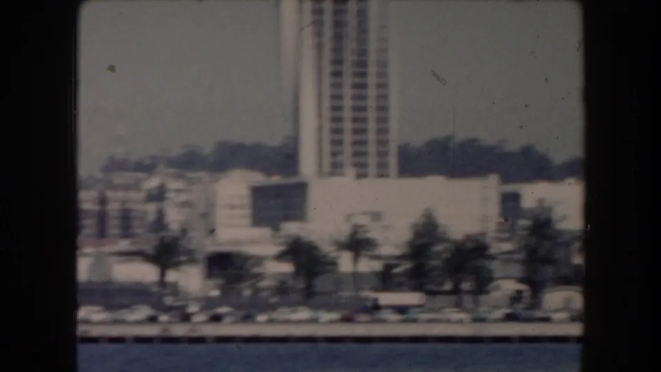 1961 California waterfront view with parked cars along the road, palm trees, low buildings, and a high-rise tower in the background.