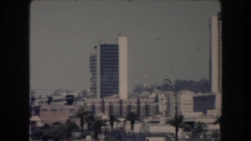 1961 California frame showing a palm-lined waterfront street, parked cars, low city buildings, and a tall rectangular tower.