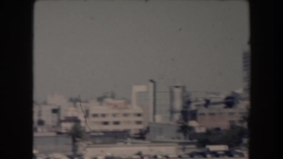 1961 California home-movie frame with distant waterfront buildings, a few taller structures, palm trees, parked cars, and open sky.