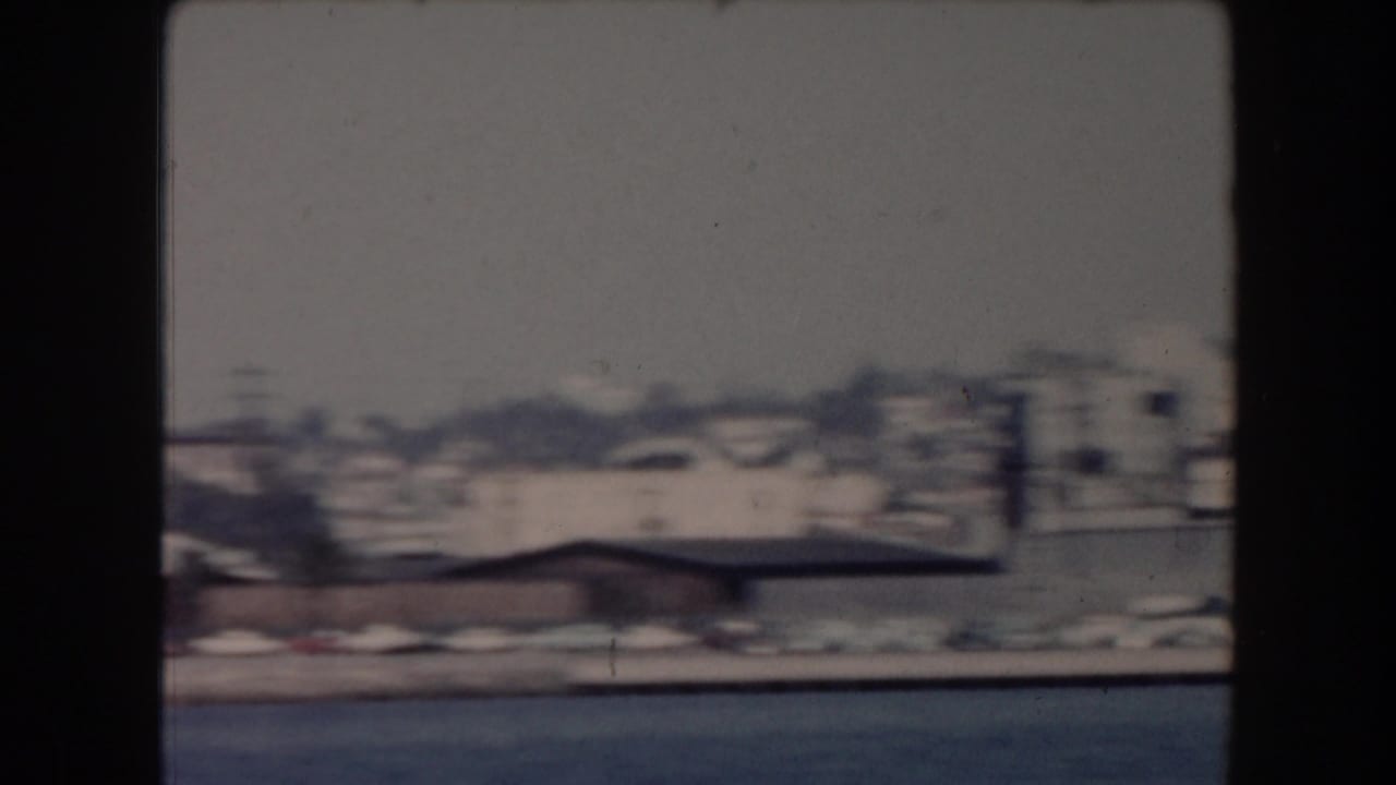1961 California home-movie frame showing a soft distant shoreline across water with low buildings and haze.