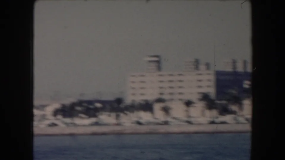1961 California waterfront frame showing an industrial-looking building with smokestacks near palms and open water.