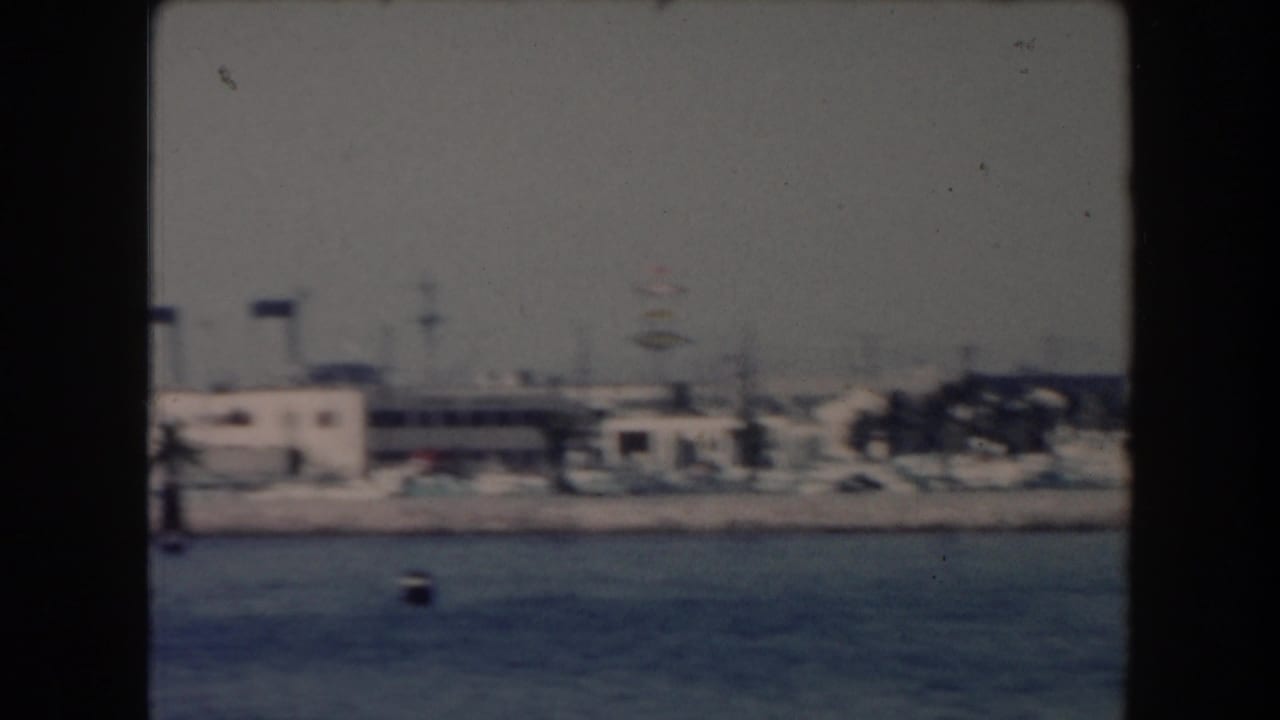1961 California home-movie frame showing a hazy waterfront with low-rise buildings, palms, and calm blue water from a boat.