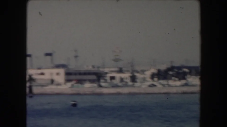 1961 California home-movie frame showing a hazy waterfront with low-rise buildings, palms, and calm blue water from a boat.