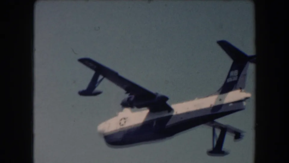 A white fuselage seaplane with dark wings and floats flies low over California coastal water in soft 8mm color.