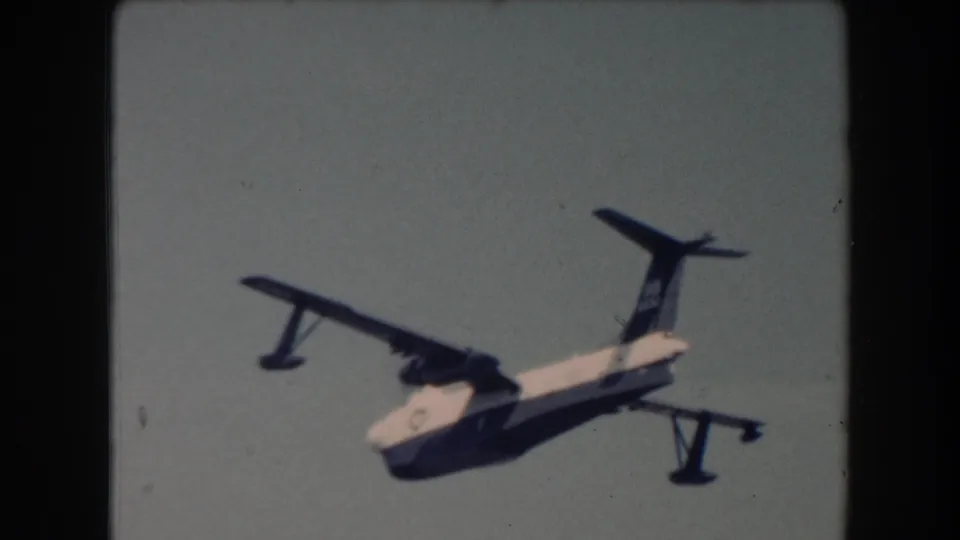 The seaplane continues across the frame over open water, with floats, dark wings, and white fuselage visible in 1961 home-movie footage.