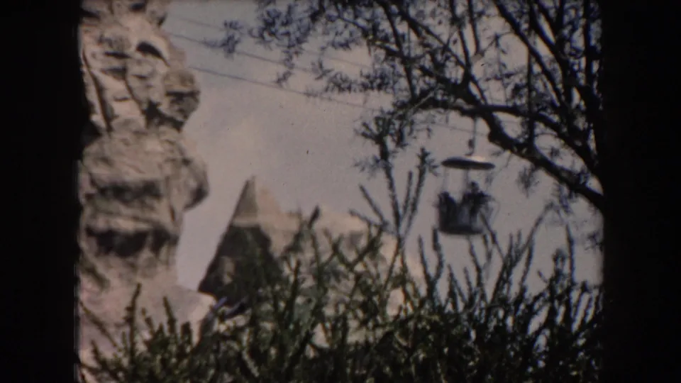 Visitor-shot 1961 California view of a cable gondola crossing in front of trees and a sculpted artificial mountain structure.