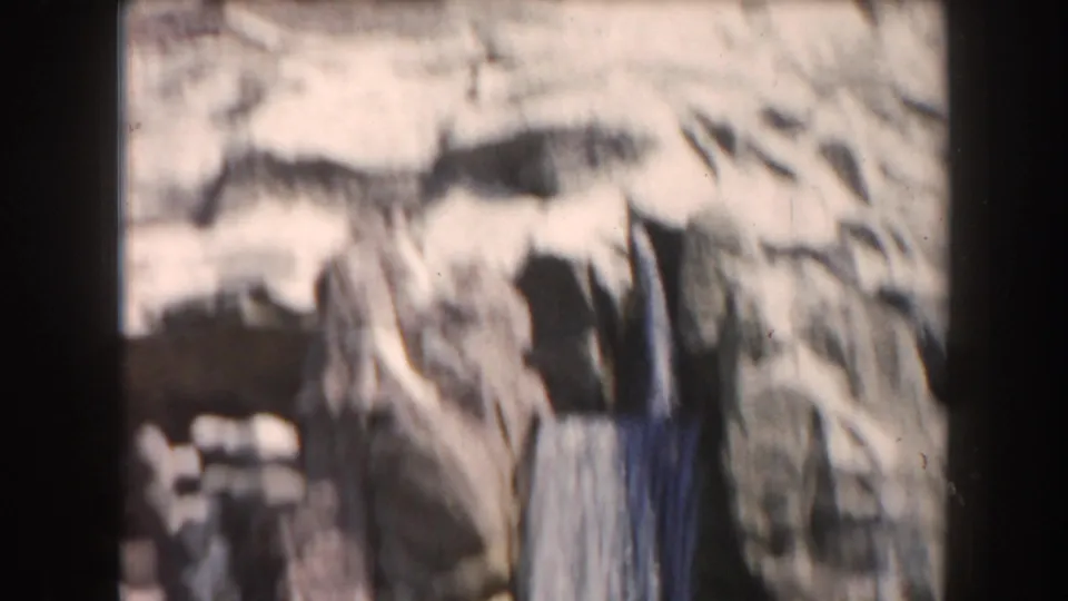 1961 California 8mm frame showing a small waterfall or water feature against pale artificial rockwork from a moving ride viewpoint.