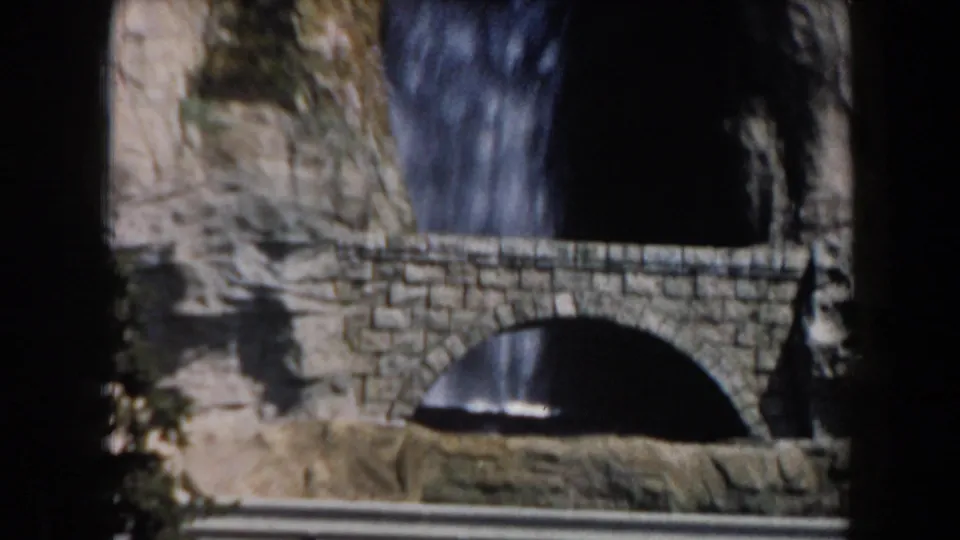 1961 California home-movie frame showing a stone arched footbridge before a waterfall and rugged rock wall.