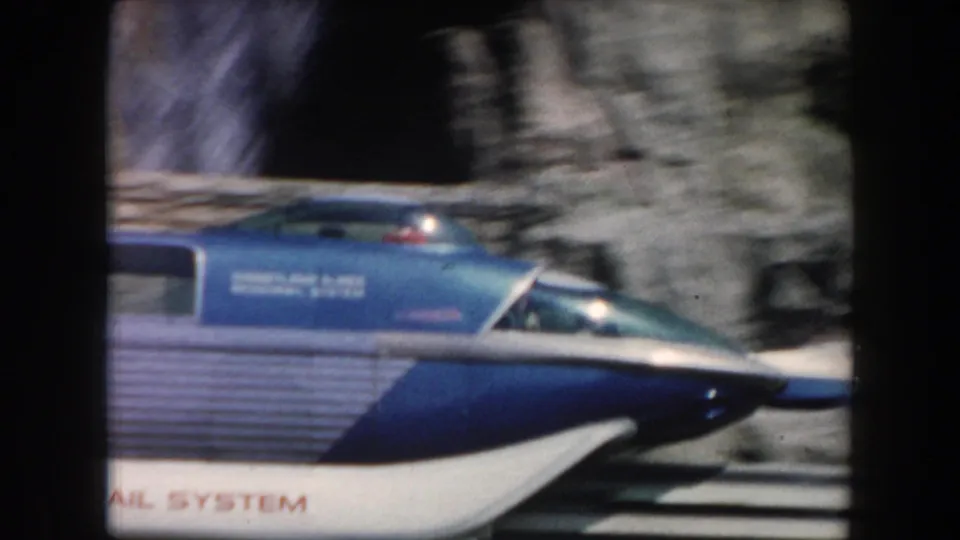 1961 California 8mm frame of a blue-and-white monorail car gliding past rocky waterfall scenery, with visible markings on the car.