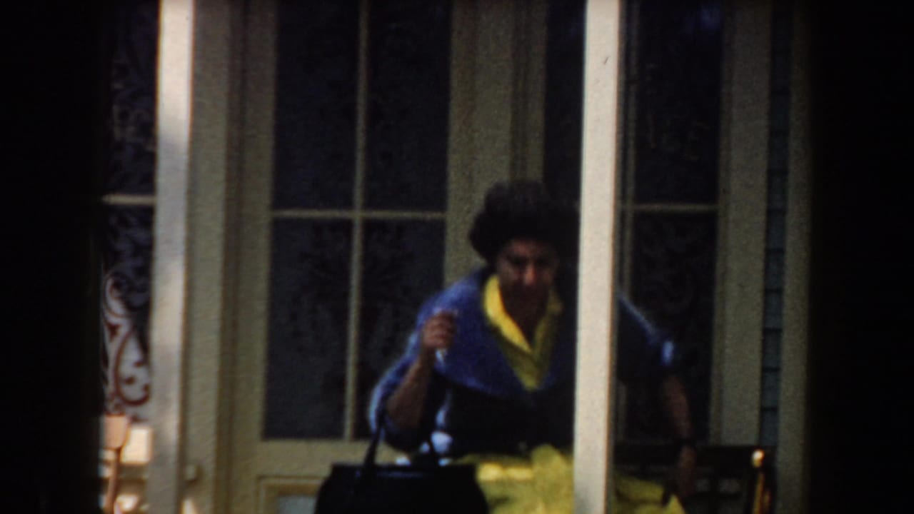 In 1961 California home-movie footage, a woman in a blue cardigan and yellow skirt approaches a porch bench near glass-paneled doors.
